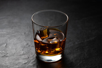 Amber whiskey with ice cubes in the old fashion glass on black stone table with studio reflection