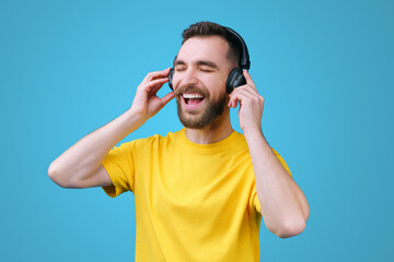 Portrait of cheerful young bearded man enjoying listening to his favourite song with closed eyes via black wireless earphones at his day-off, wearing casual yellow t-shirt, isolated on blue background