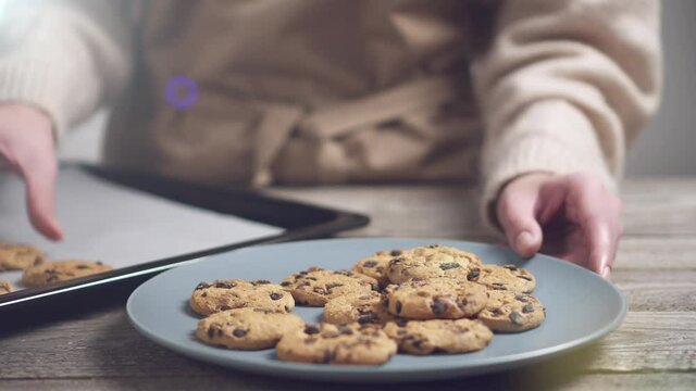 Homemade chocolate chip cookies on a baking sheet lined with parchment paper. A girl in a beige sweater puts cookies on a white plate. A slide of chocolate chip cookies on a wooden kitchen table.