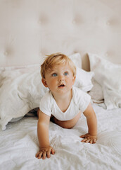 Little blond boy in a white bodysuit sitting on the bed.