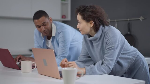 Side angle view of concentrated Caucasian woman and African American man typing on laptop keyboard in slow motion smiling to each other. Happy young couple working online from home office