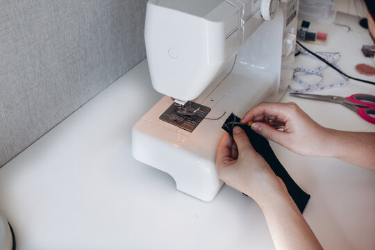 Side View Of The Sewing Process On The Sewing Machine, Hands In The Process Of Sewing Black Fabric, White Background. Hobby And Seamstress Concept