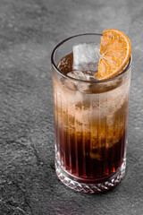 Cocktail in glass with cola, alcohol, ice, decorated dried orange on gray background.