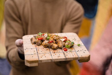 Fried mussels with chili, garlic, sesame, herbs, on square wooden board. Serving the dish by the waiter.