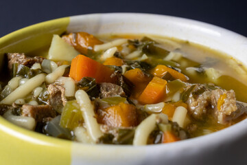 Soup in a yellow and white bowl positioned on a dark black background containing potatoes, carrots, pasta, beef and green seasoning. In detail each of the elements in pieces.