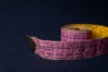 Measuring tape in pink and yellow colors with a metal tip on a dark black background with left to right illumination. Reference to weight loss, measurements in general, healthy living and sewing.