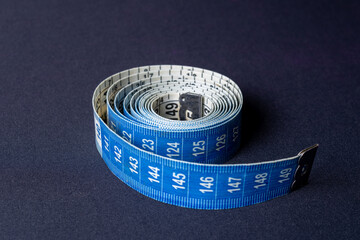 Blue and white measuring tape with a metal tip on a dark black background with left-to-right illumination. Reference to the weight loss process, measurements in general, healthy living and sewing.