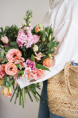 Celebrating spring. Close up shot of young stylish female in casual wear with wicker bag holding gorgeous big bouquet with fresh flowers in bloom while standing against grey background, vertical shot