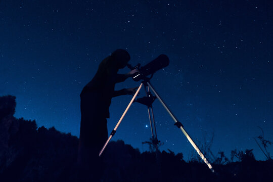 Dark silhouette of a sky watcher of the starry sky in a telescope. Woman observer watching night sky in a reflector. Image may contain noise grain due to high ISO range when shooting very dark scenes.