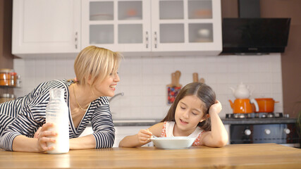 The concept of breakfast in the kitchen. Young mother pours milk on the plate of her cute little...