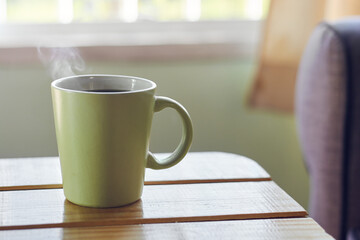 A view of a green ceramic coffee cup with steam on wooden rustic table in cozy living room interior against window light.