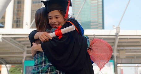 Graduation Day Celebration, Excited Graduate Student in Gown and Cap with Diploma Hugs his Mother after Graduation Ceremony. Asian mom embraces daughter with joy on graduation day and successful.