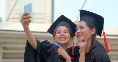 Best friends hugging warmly and laughing, graduation gown use mobile phone to selfie together in big city. Concept of celebration and congratulation after success of education in high level.