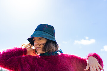 young latina woman posing with her hands in the air, against the light, wearing a bucket hat with a red sweater, blue eyes, blue eyes. - obrazy, fototapety, plakaty