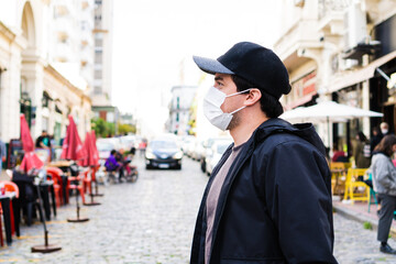 side shot of a man wearing a surgical face mask while walking down a street full of restaurants.