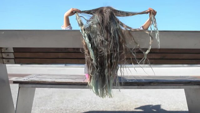 Brunette preteen girl with blue color highlights sitting on the bench on the bridge and brushing her hair with hands in the wind, sunny summer day, back view, natural beauty