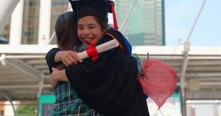 Graduation Day Celebration, Excited Graduate Student in Gown and Cap with Diploma Hugs his Mother after Graduation Ceremony. Asian mom embraces daughter with joy on graduation day and successful.