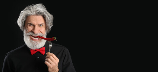 Senior man eating chili pepper on dark background