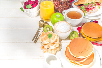 Various traditional breakfast food - fried eggs with bacon, muesli, oats, waffles, pancakes, burger, croissants, fruit berry, coffee, tea and orange juice, white table background copy space top view