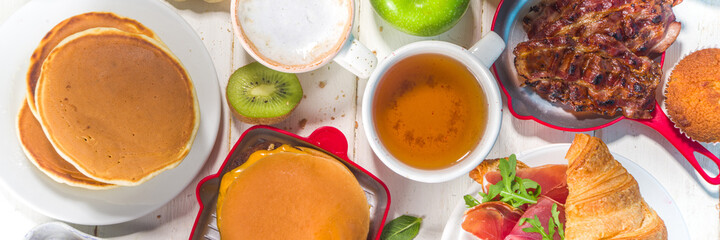 Various traditional breakfast food - fried eggs with bacon, muesli, oats, waffles, pancakes, burger, croissants, fruit berry, coffee, tea and orange juice, white table background copy space top view