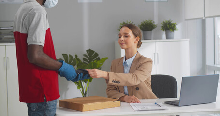 Obraz premium Deliveryman in mask and gloves giving pizza box to female office worker paying with credit card