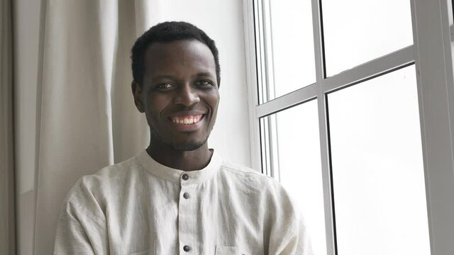 Handsome young African-American man in linen shirt turns head and smiles to camera sitting near large window in room closeup