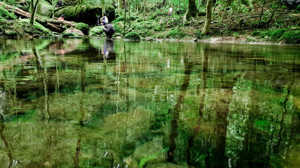 Traveller and photographer are taking the photo of waterfall, Clear river with green rocks and trees above the water surface, Nature leaves shadow over the river in the dark forest