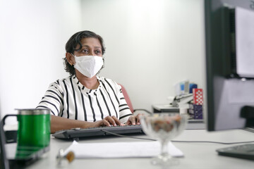 Fototapeta na wymiar Asian Indian businesswoman with medical face mask typing on her computer keyboard in the office