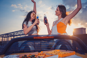Two young girls smiling, cheering with soda in glass bottles, posing in yellow car cabrio with french fries and pizza on trunk. Fast food. Copy space