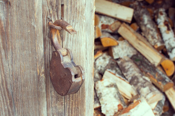 Old barn door lock hanging on a loop against the background of old textured wooden walls of boards and dried firewood from birch wood.
