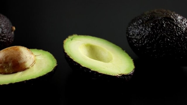 Halved avocado with a stone and two whole ripe avocados on a black background, 4K UHD dolly side pan 