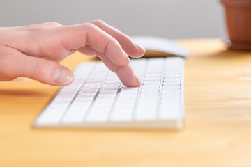An image of a woman's hand is typing on a white keyboard on a wooden table. Selective focus
