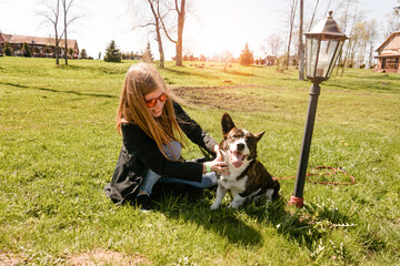 Naklejka na ściany i meble Young woman in red sunglasses plays with her corgi on a summer green lawn. Sunny day. Happy pet and owner