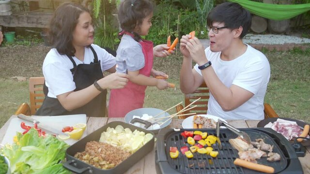 Happy Asian family enjoy and having fun weekend activity together at home. Father and mother with little daughter cooking grilled meat and vegetable barbecue on cooking pan in garden together at home.