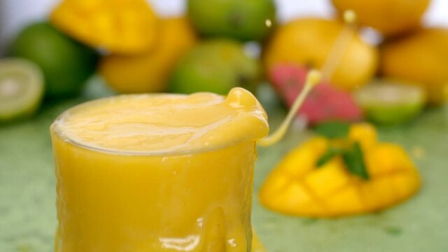 Big ice cubes falling / splashing into a glass full of refreshing mango smoothie - Summer drink India. Closeup shot of sliced mangoes garnished with mint leaves and citrus lemon halves against blur...