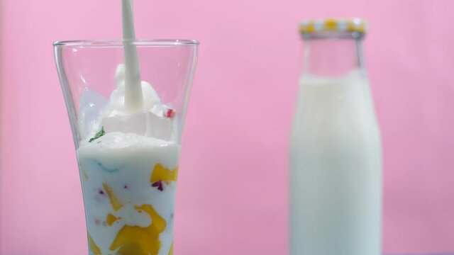 Milk pouring in a glass with mango pieces  ice cubes  tutti frutti  and mint leaves - Summer drink India. Closeup shot of a fresh bottle of milk placed with a healthy tropical mango / Aam fruit smo...