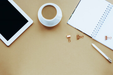 Overhead workplace with tablet computer, notepad, pen, coffee cup on beige background. Top view, flat lay, copy space