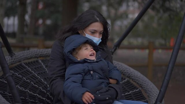 Mother wearing covid-19 face mask in swing with toddler son, mom bonding during covid