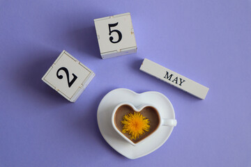 Calendar for May 25: cubes with the number 25, the name of the month of May in English, a cup of coffee in the shape of a heart with a dandelion flower, top view