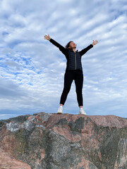 A young woman in sportswear stands on the top of a mountain with her arms raised up against the background of the sky.