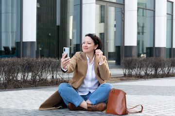 An adult woman sits on the sidewalk and speaks via video link with a joyful expression on her face.