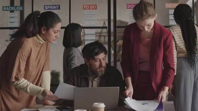 Steadicam of bearded Caucasian man sitting by desk using portable computer, two diverse female colleagues standing on his sides, showing business papers in busy office