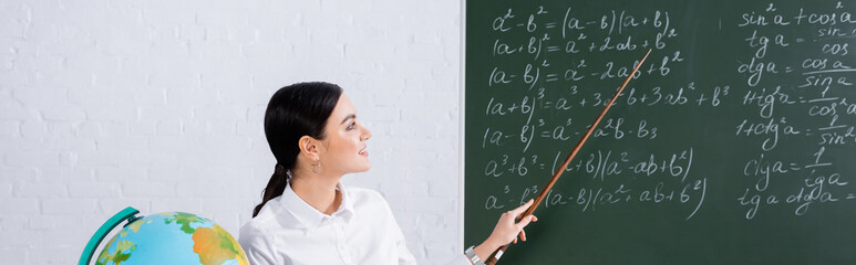 Smiling teacher pointing at mathematic equations on chalkboard near globe, banner