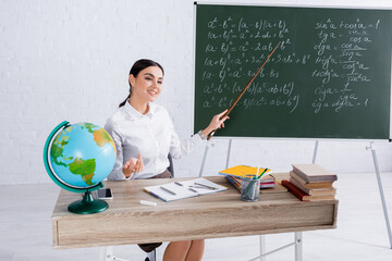 Cheerful teacher with pointer sitting near chalkboard and globe during lesson