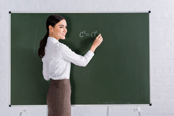 Cheerful teacher with chalk writing mathematic equation on chalkboard