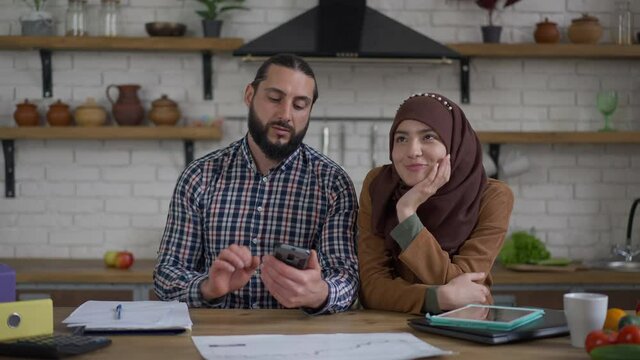 Successful handsome Middle Eastern husband discussing business strategy wife beautiful charming wife in hijab. Portrait of positive smiling couple sitting in kitchen in home office indoors talking