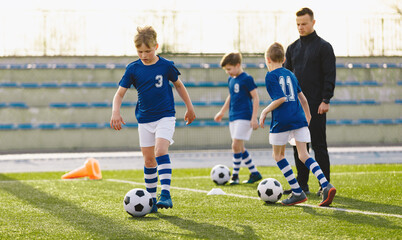 Young sports boys in blue sportswear running and kicking a ball on the pitch. The soccer youth team plays football training in the summer. Sports activities for kids. Soccer coach playing 