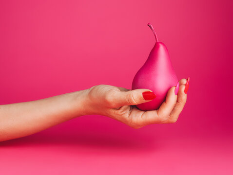 Woman hand holding pink painted pear against matching pink background. Creative fruit food concept.