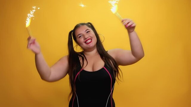 Happy holiday. Funny plus size woman. Party time. Body positive. Holiday celebration. Cheerful obese lady in tank shirt dancing with firework sparklers in hands isolated yellow.