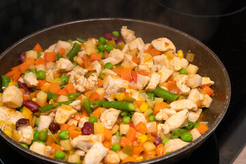 Pieces of white meat, a variety of vegetables are fried in a frying pan on an induction stove